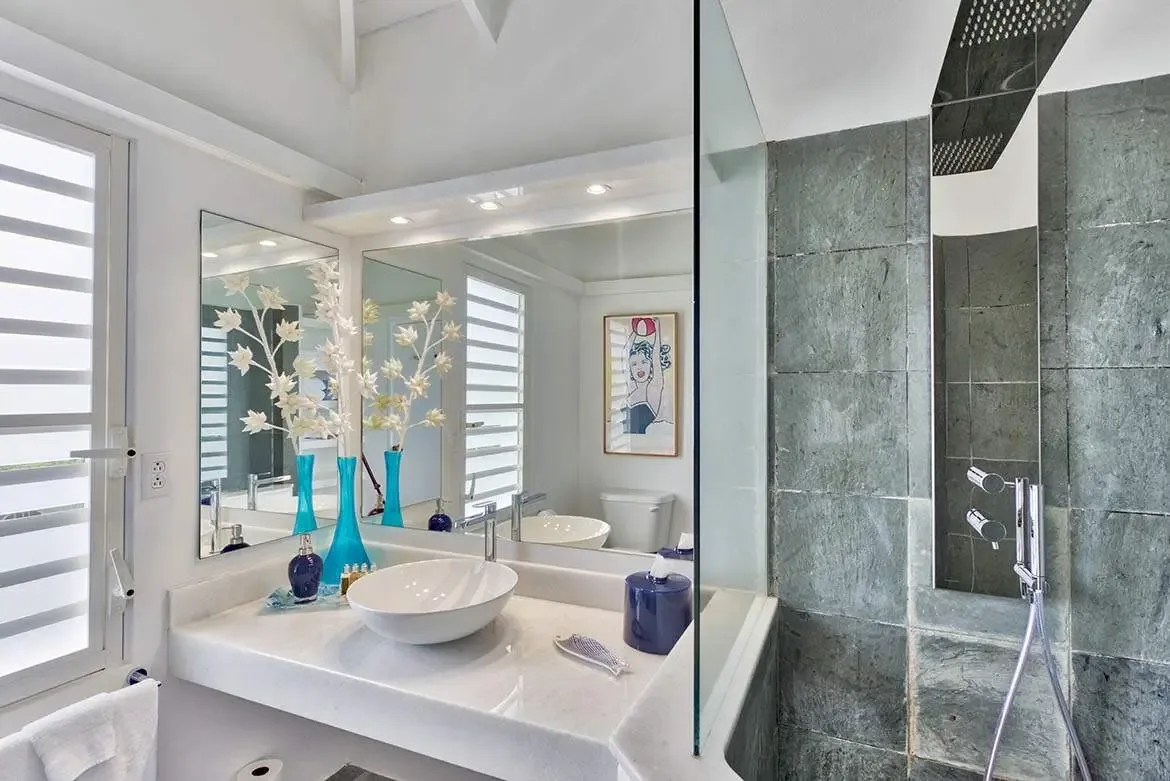 A white marble vanity with a vessel sink holds blue glass vases with white floral branches beside a frameless mirror, where a glass-enclosed shower with slate walls and a rain showerhead adjoins.