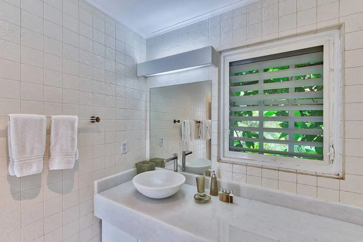 A white marble vanity with a vessel sink and chrome faucet sits beneath a frameless mirror, where jalousie windows reveal lush tropical foliage and gold-toned accessories add warm accents.