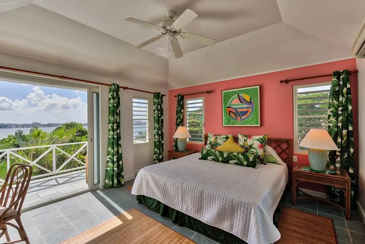 A bamboo headboard with floral and palm-leaf pillows anchors a coral-pink accent wall with abstract art, where rattan nightstands and green curtains frame a balcony with an ocean view.