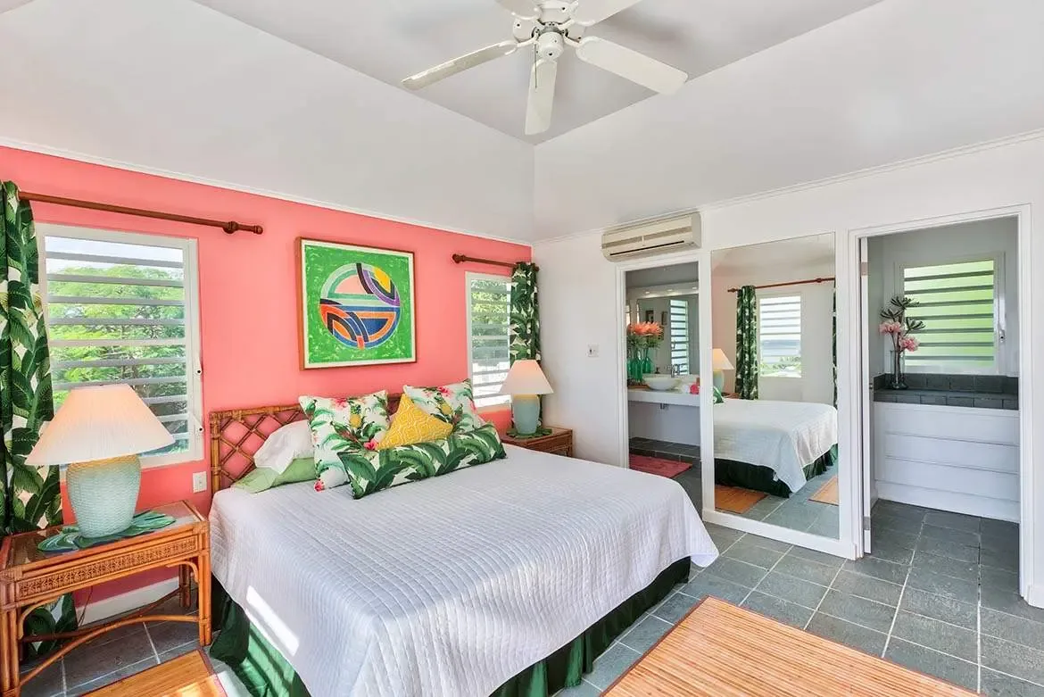 A white quilted bed with tropical-leaf and yellow pillows sits before a coral accent wall with abstract green art, where a mirrored closet door and an open doorway to an en-suite bathroom reveal louvered windows.
