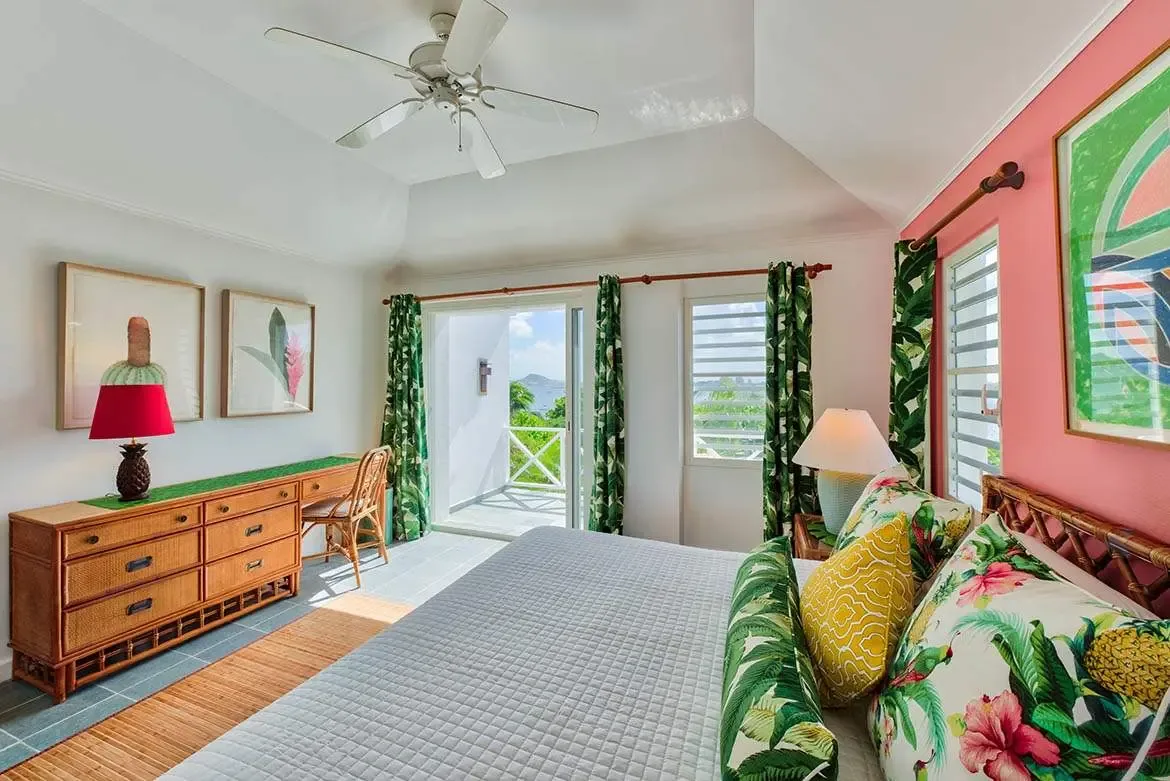 A white quilted bed with hibiscus and pineapple pillows sits before a salmon-pink accent wall with botanical prints, where a rattan desk and a balcony overlook lush tropical foliage and the blue ocean.