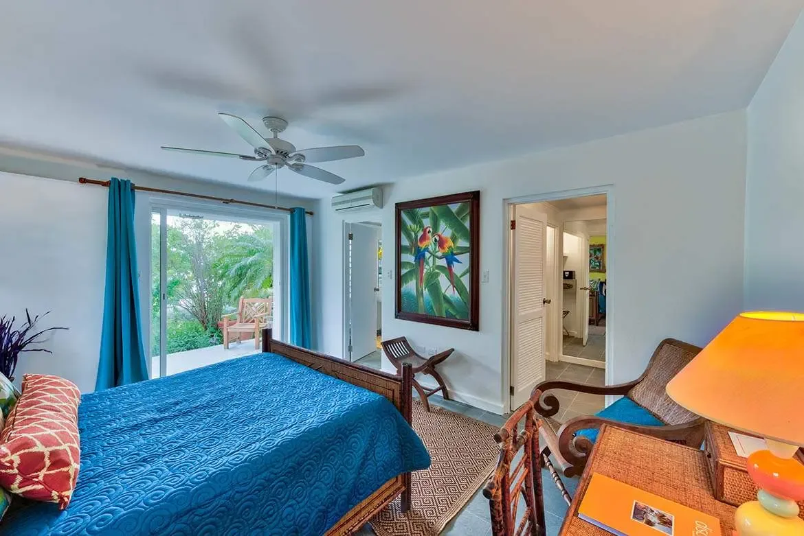 A dark wood bed with a teal quilt and a red bolster sits beside a painting of colorful parrots, where a wooden armchair with a blue cushion and an orange lamp face glass doors opening to tropical greenery.