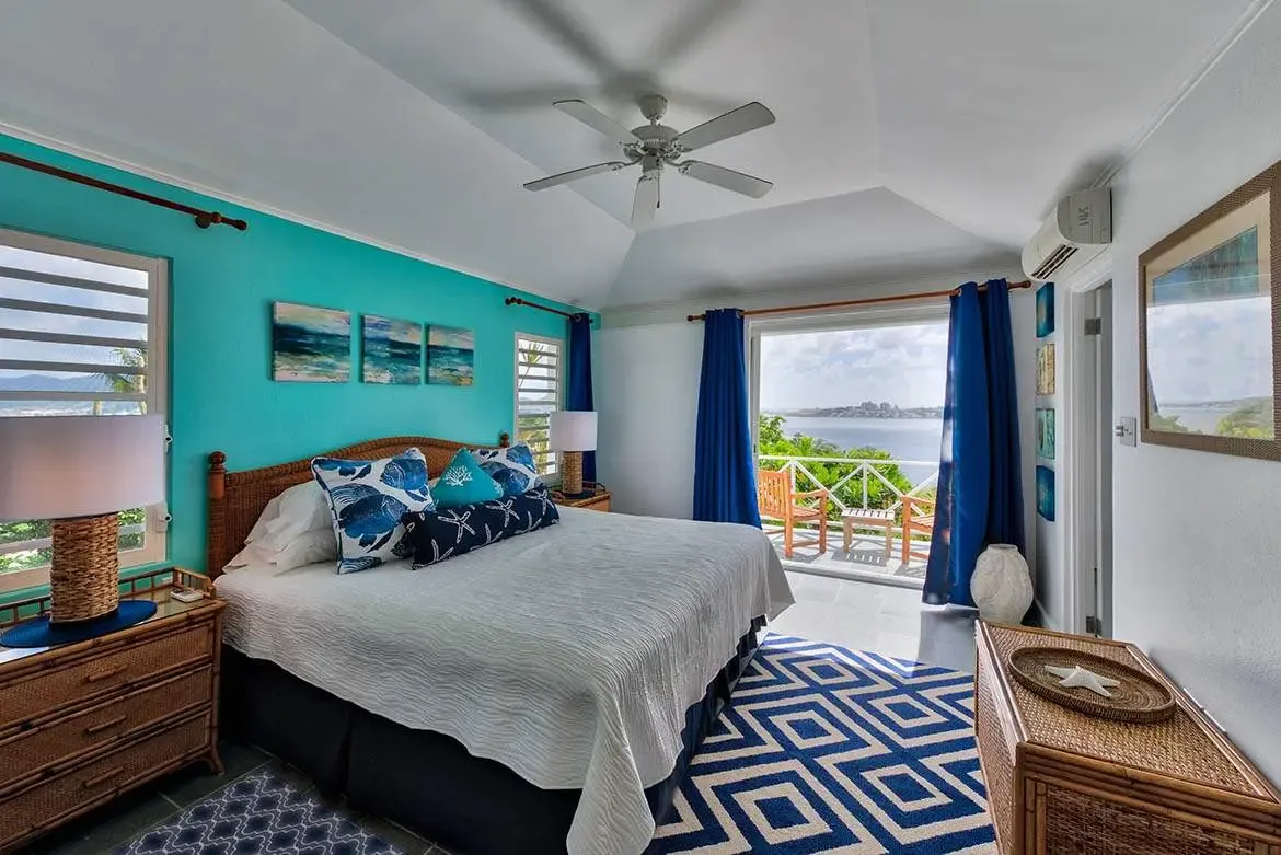 A rattan headboard with starfish and botanical pillows anchors a turquoise accent wall with ocean-horizon canvases, where navy curtains frame a balcony with sweeping turquoise ocean views.