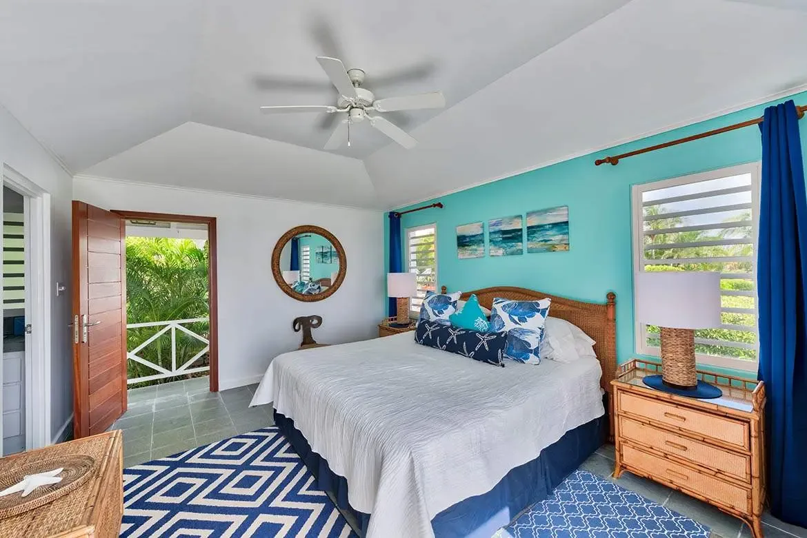 A wicker headboard with blue floral and turquoise pillows sits before a turquoise accent wall with ocean canvases, where a mirrored closet and a balcony with tropical foliage reflect the room's coastal charm.