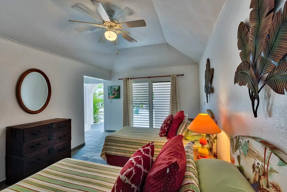 Two twin beds with green striped coverlets and red pillows sit beneath large metal leaf sculptures on a white wall, where an orange-shaded lamp and a wicker dresser stand beside glass doors with greenery beyond.