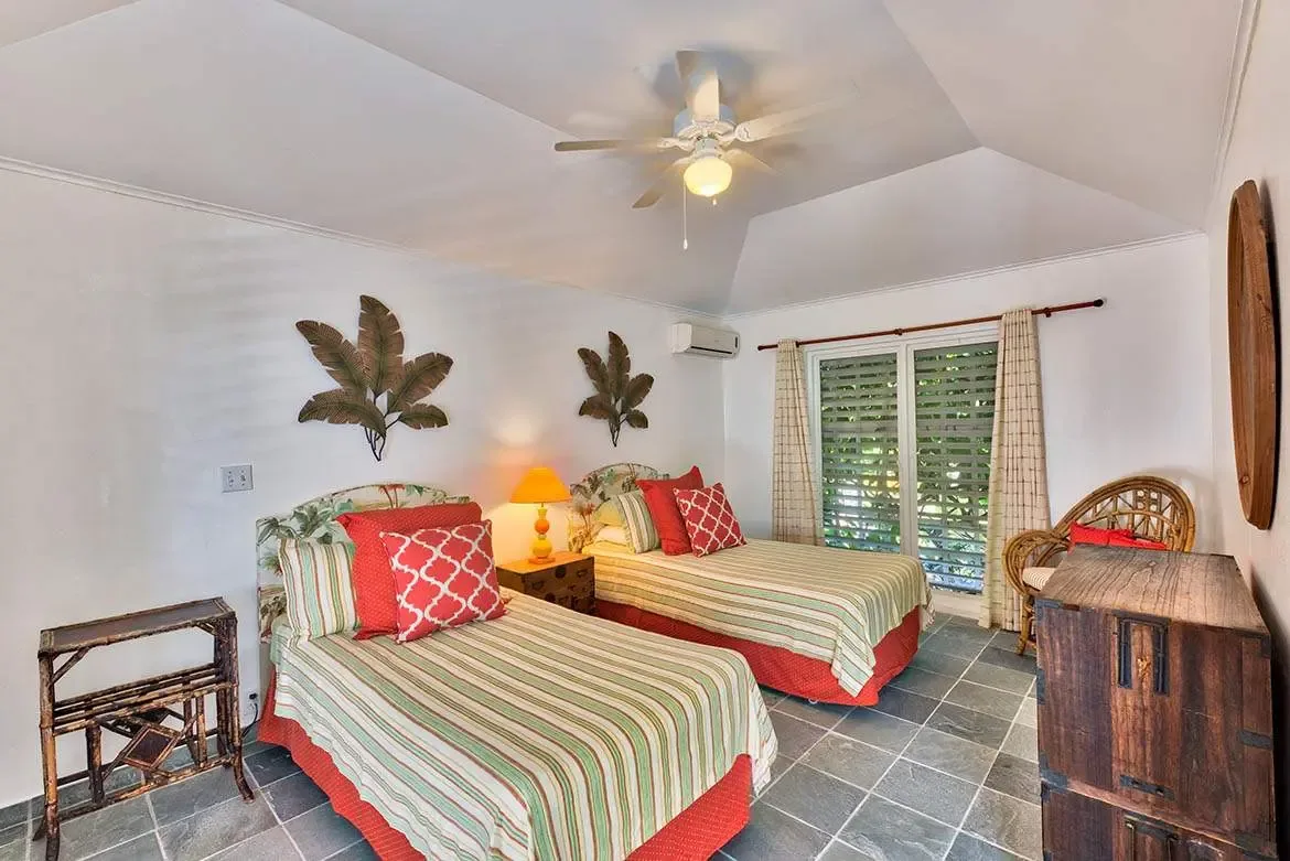 Two twin beds with striped coverlets and red skirts sit beneath arched tropical-print headboards and dark leaf sculptures on a white wall, where a vaulted ceiling and glass doors reveal lush greenery outside.