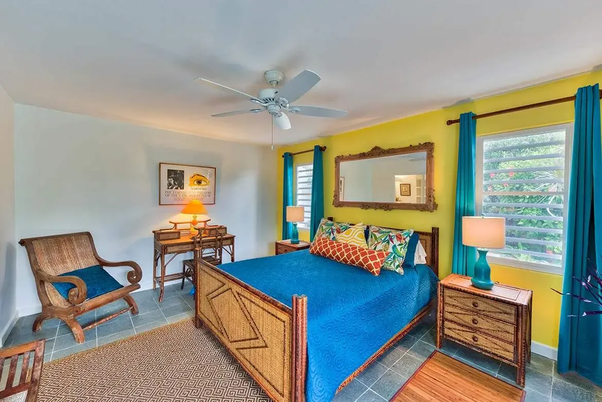 A bamboo bed with a teal quilt and tropical-leaf pillows sits before a yellow accent wall with an ornate gold-framed mirror, where a rattan desk with an orange lamp and turquoise curtains frame a garden view.