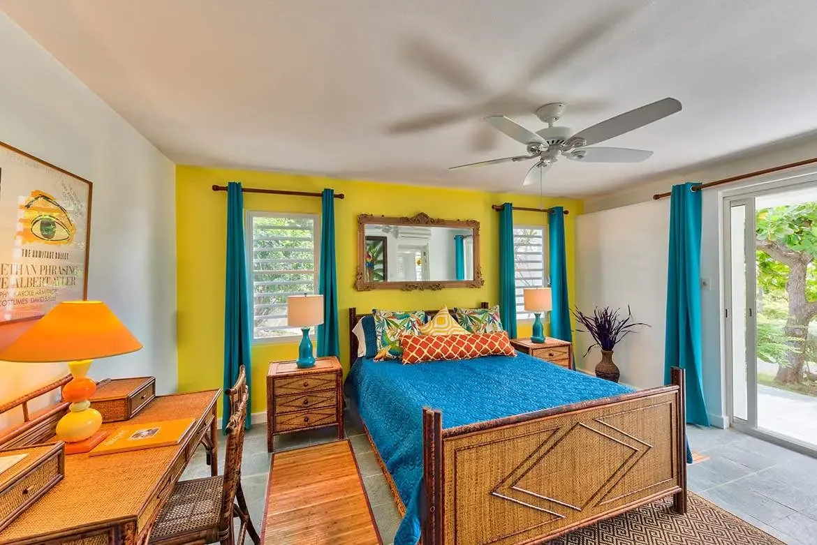 A bamboo bed with a teal quilt and orange geometric bolster sits before a yellow wall with a gold mirror, where a rattan desk with an orange lamp and purple branches in a vase flank a balcony door.