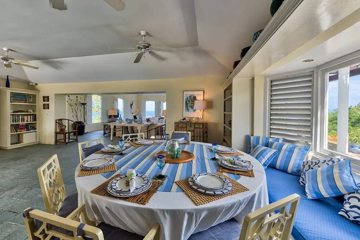 A round table with blue-and-white striped settings and geometric-back chairs sits beneath a vaulted ceiling, where a blue-cushioned window seat and louvered shutters open to an ocean view.