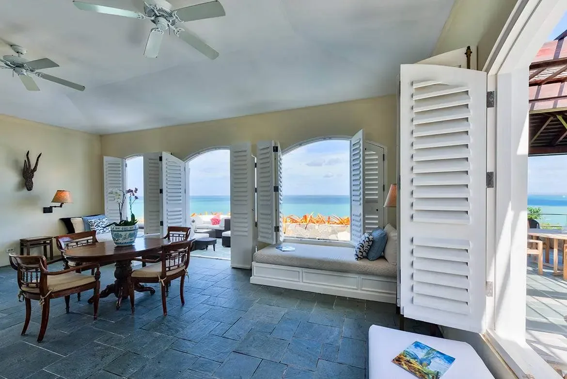 An oval dark wood table with an orchid centerpiece and woven-back chairs sits on slate tiles, where arched shutters open wide to a turquoise ocean and a window seat invites lingering.