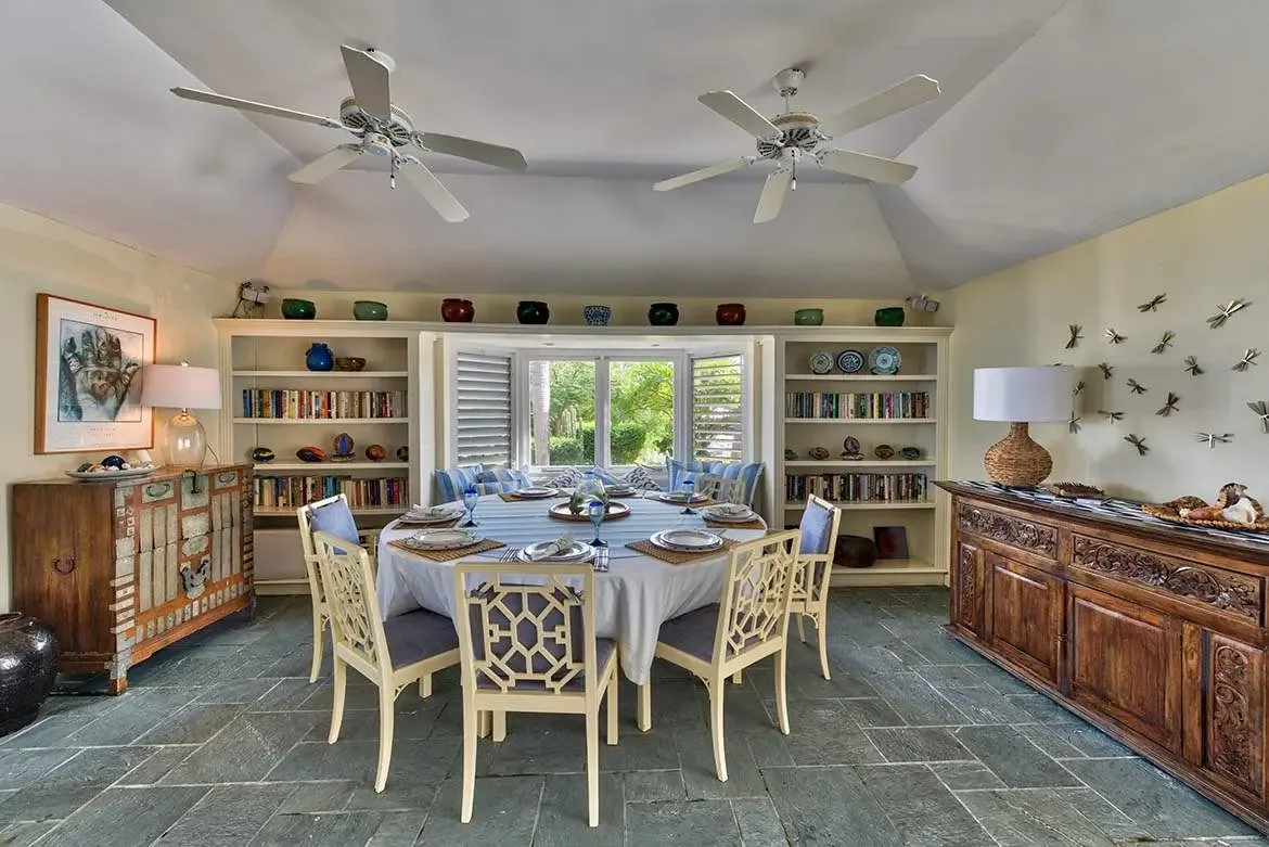 A round dining table with blue-and-white settings and six geometric-back chairs fills a vaulted room with yellow walls, where built-in bookshelves and metallic dragonfly sculptures frame a window seat with green foliage beyond.