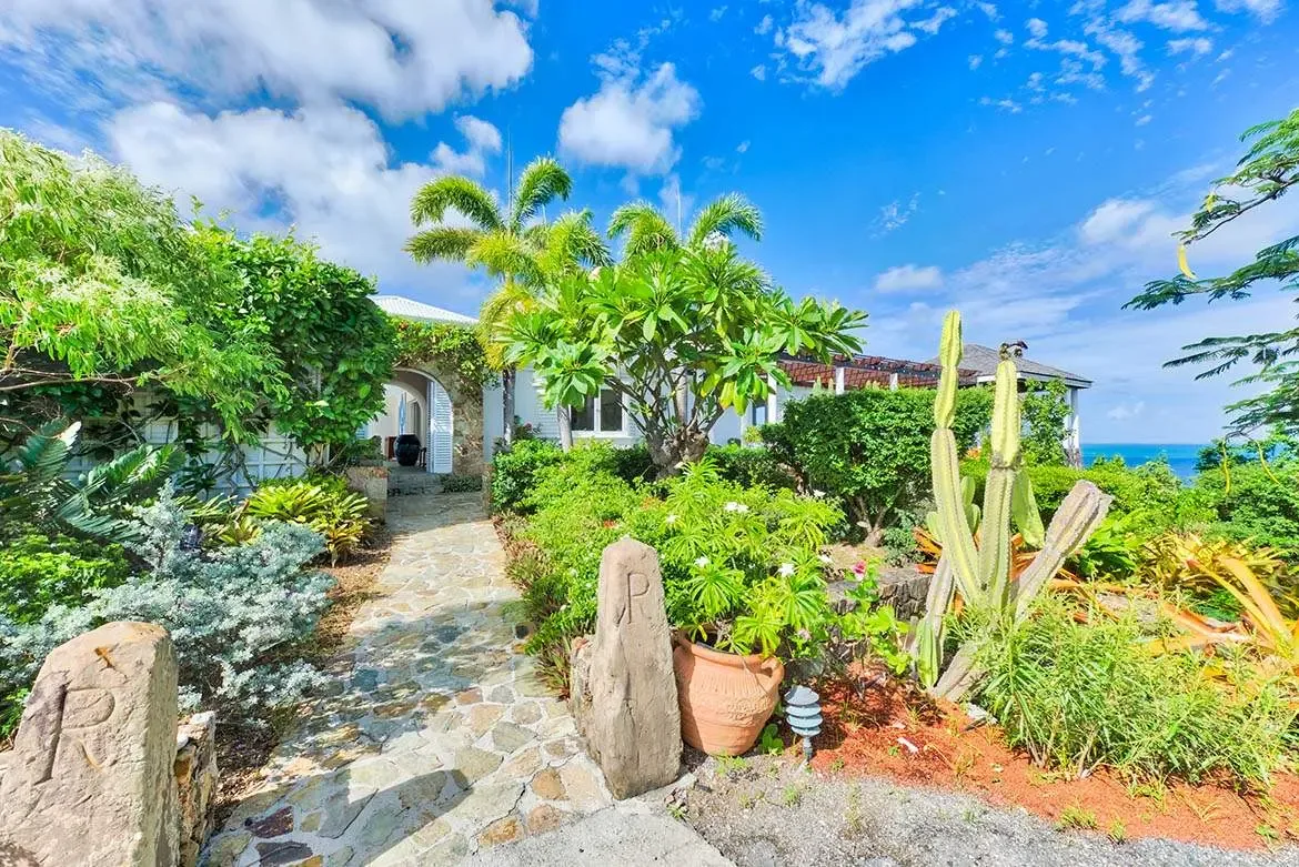 Stone pillars with carved "R" motifs flank a path of irregular slate slabs through lush tropical gardens, where palm trees and blooming bromeliads frame a white villa with terracotta roof and distant ocean view.