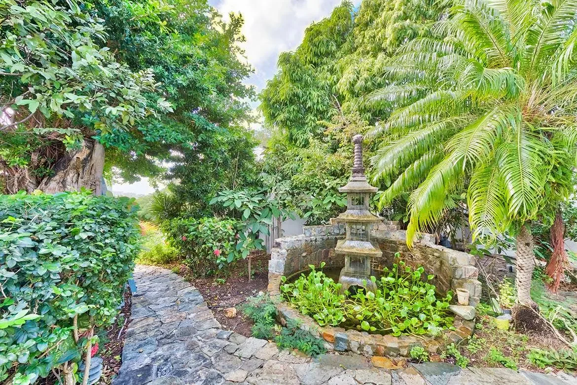 A multi-tiered stone pagoda stands in a circular basin of green lily pads, where a winding slate path leads through dense tropical foliage toward a sunlit hillside beyond.