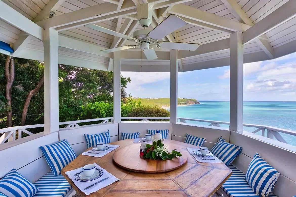 A hexagonal wooden table with a lazy susan and blue-and-white settings sits on built-in benches with navy-striped cushions inside a white gazebo, where the turquoise ocean stretches to a distant shoreline.