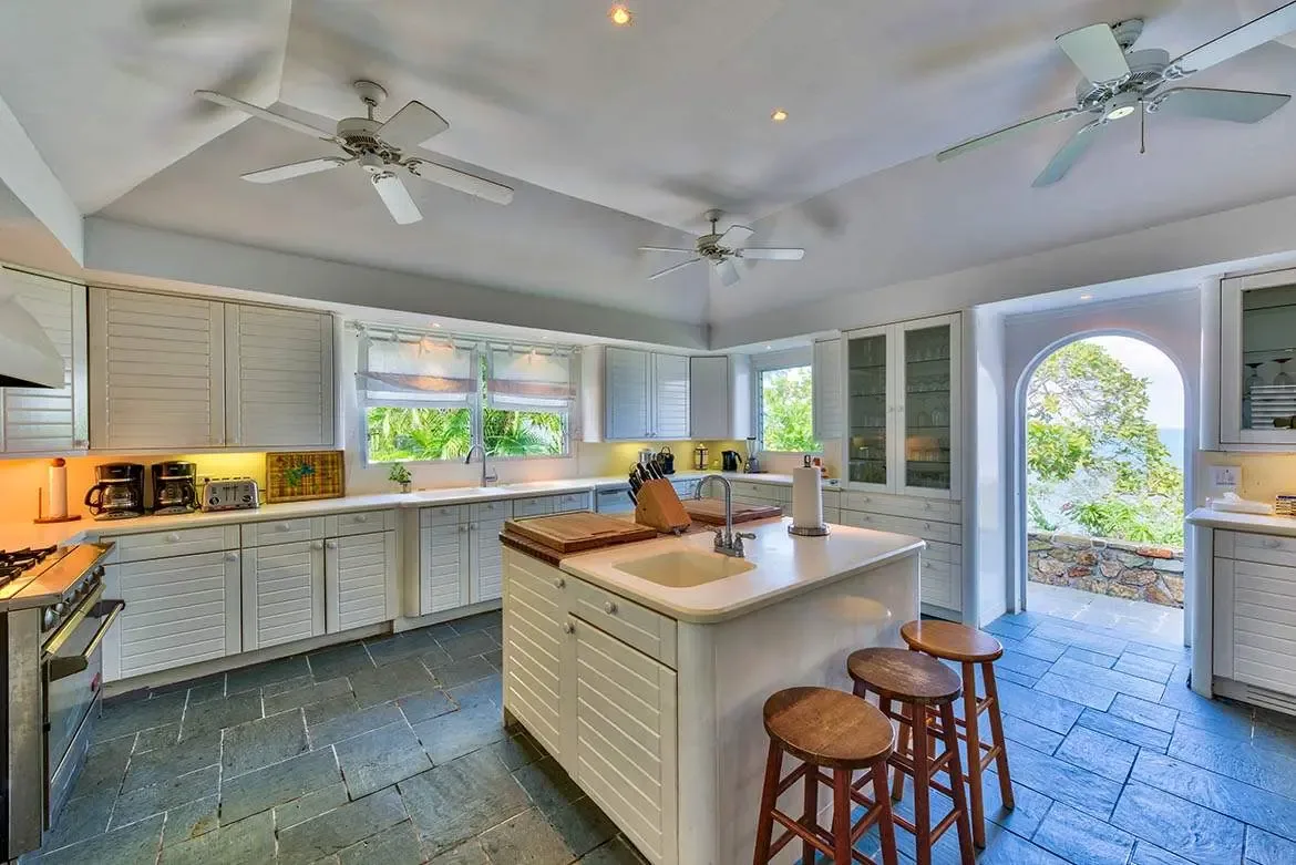 White louvered cabinetry and a butcher-block island with bar stools fill a vaulted kitchen beneath three ceiling fans, where an arched doorway opens to lush tropical greenery and a glimpse of blue ocean.