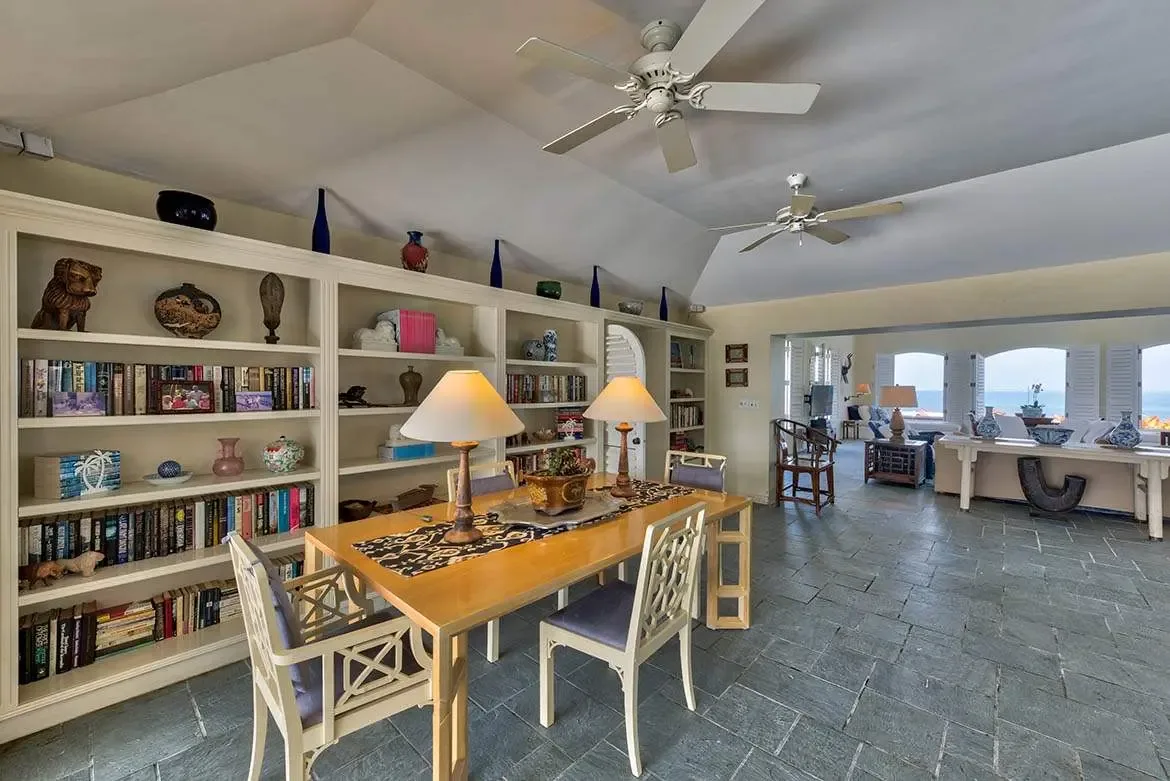 Floor-to-ceiling white bookshelves filled with books and blue glass bottles line a vaulted room, where a glass table with lattice chairs sits beside a living area with an ocean panorama beyond arched windows.