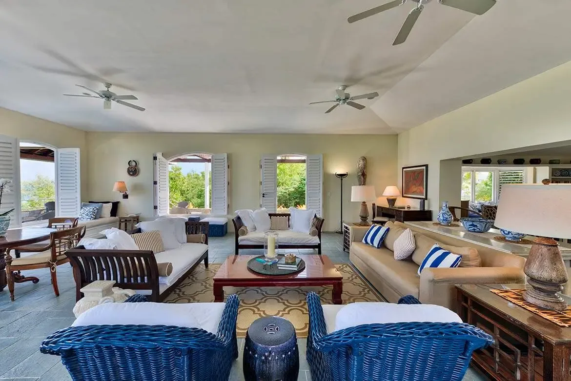 Royal blue wicker armchairs and a camel sectional with blue-striped pillows anchor a vaulted living room, where arched shutters reveal the ocean and a dining area sits beyond a console with blue ceramics.