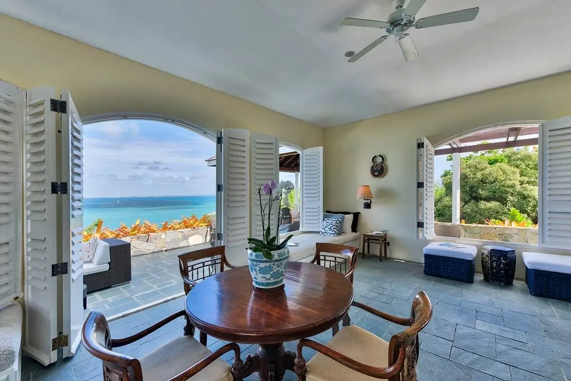 A round table with a purple orchid sits beside navy ottomans and a window daybed with blue pillows, where two arched openings frame the turquoise ocean and lush tropical gardens.