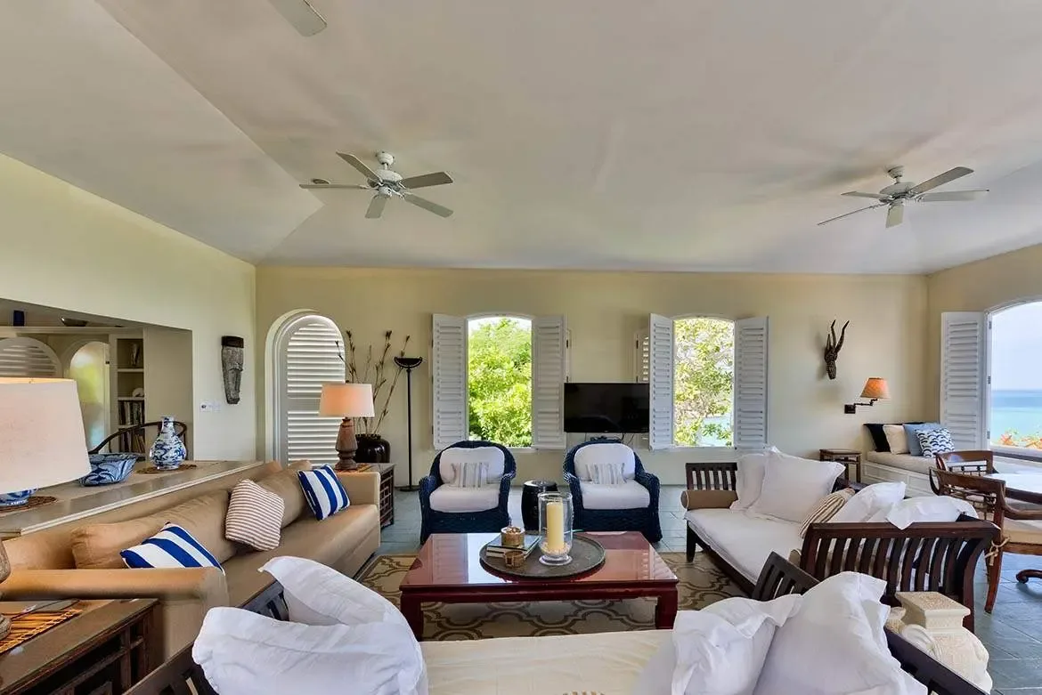 A tan sectional with navy pillows faces dark wood armchairs across a patterned rug, where three arched windows with louvered shutters reveal lush greenery and a distant turquoise ocean.