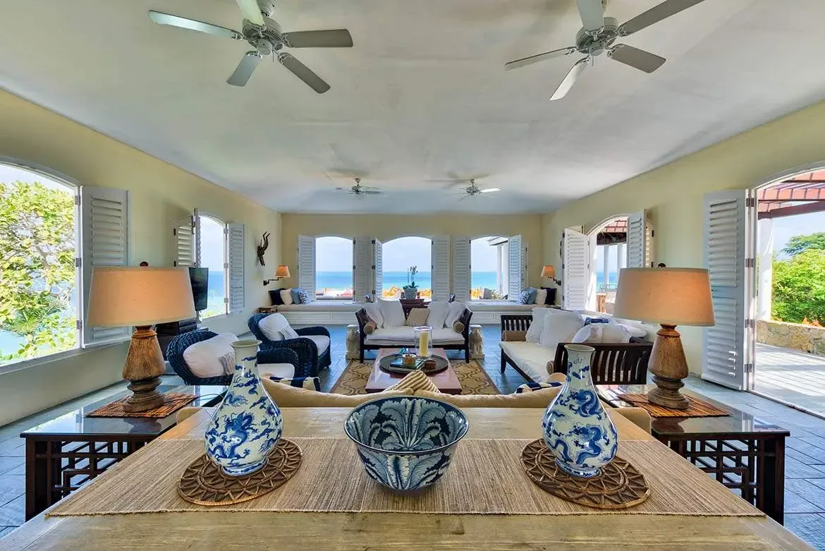 Two white sofas and navy armchairs sit on a patterned rug around a wooden coffee table beneath four ceiling fans, where blue-and-white ginger jars and arched shutters frame sweeping turquoise ocean views.