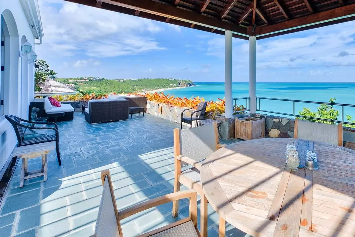 A weathered wooden dining table sits under a beamed ceiling between white columns, where wicker sofas with pink pillows and slate tiles lead to a stone wall topped with orange bromeliads overlooking a turquoise bay.