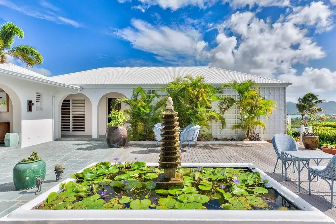 A square lily pond with purple blooms and a stone pagoda fountain anchors a wooden deck, where light-blue wicker chairs sit beside a white villa with arched openings and louvered shutters overlooking green hills.
