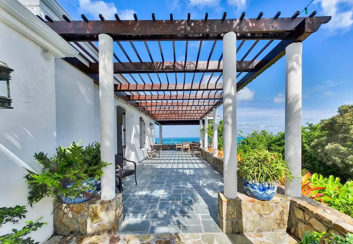 A white-columned pergola casts geometric shadows on slate tiles, where blue-and-white planters overflow with ferns and a dark armchair faces the turquoise ocean beyond a stone railing.