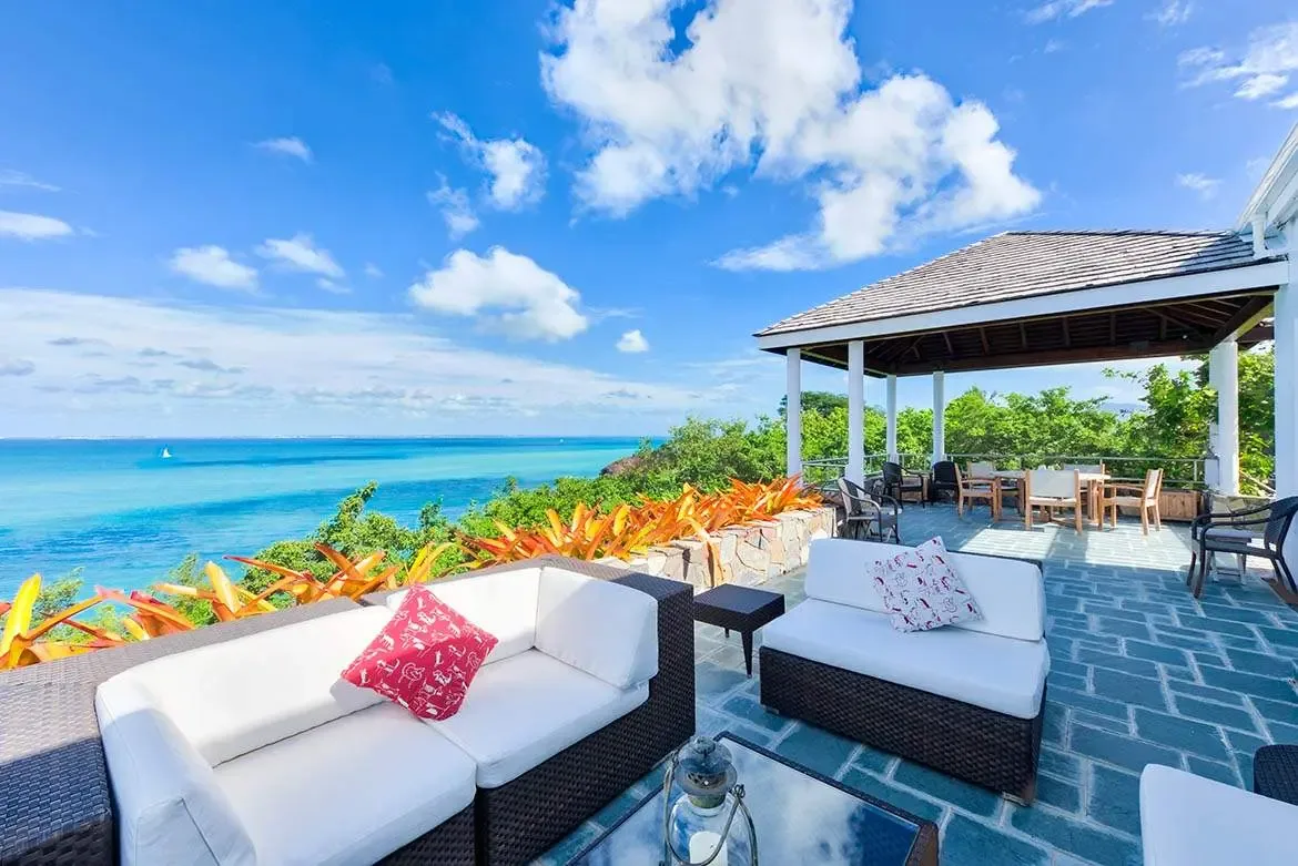 White-cushioned wicker sofas and a red accent pillow sit on a slate terrace beside orange bromeliads, where a covered dining gazebo overlooks a turquoise bay with a lone sailboat on the horizon.