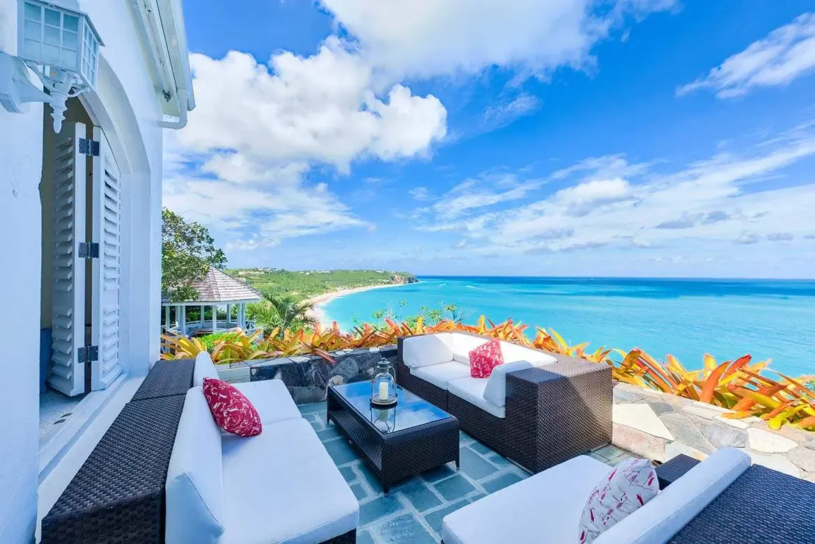Dark wicker sofas with white and red pillows sit on a stone terrace beside orange bromeliads, where a white gazebo overlooks a sweeping crescent beach and turquoise bay beneath scattered clouds.
