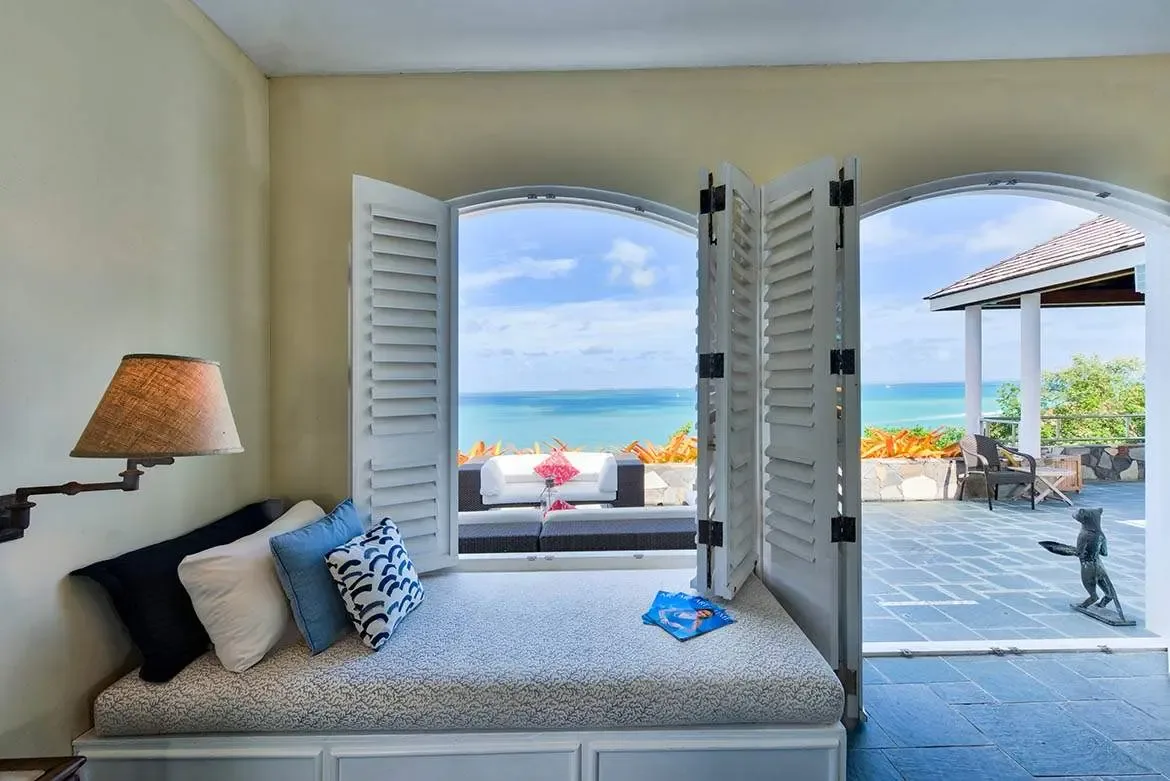A window daybed with navy and blue pillows sits beneath a swing-arm lamp beside arched shutters, where a stone terrace with orange bromeliads overlooks a turquoise ocean stretching to the horizon.