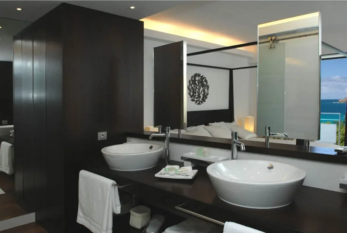 Two white vessel sinks gleam on a dark polished countertop with modern chrome faucets and a warm light strip above. The mirror reflects the bedroom beyond and the turquoise ocean outside—where even your morning routine comes with a Caribbean view.