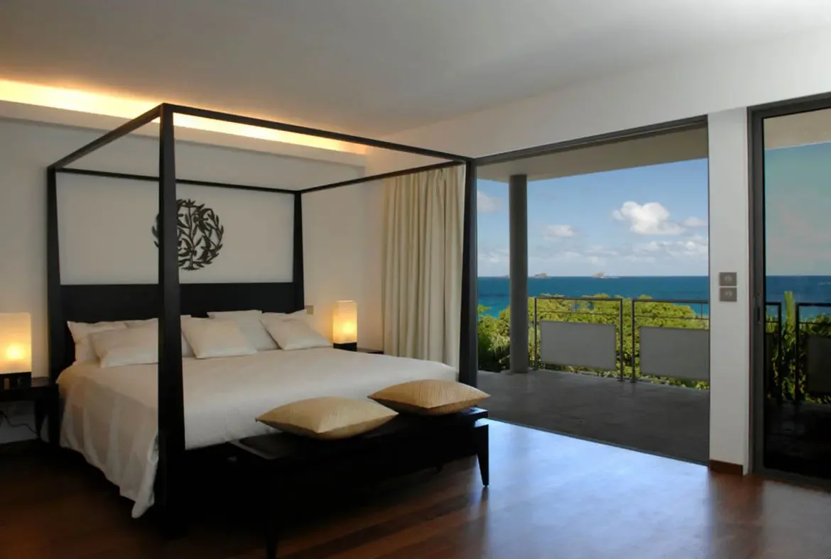 A black four-poster bed with crisp white linens and gold throw pillows on a bench at the foot anchors this elegant bedroom. Floor-to-ceiling glass doors open to a terrace overlooking the blue ocean and distant islands—modern luxury meets Caribbean panorama.