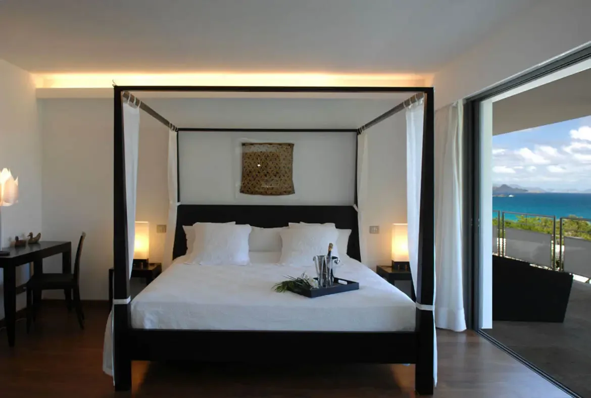 A black four-poster bed with white linens awaits with a champagne bucket and glasses on a bedside tray—romance ready. Floor-to-ceiling glass doors open to a terrace where the turquoise ocean stretches to distant islands beneath a bright Caribbean sky.
