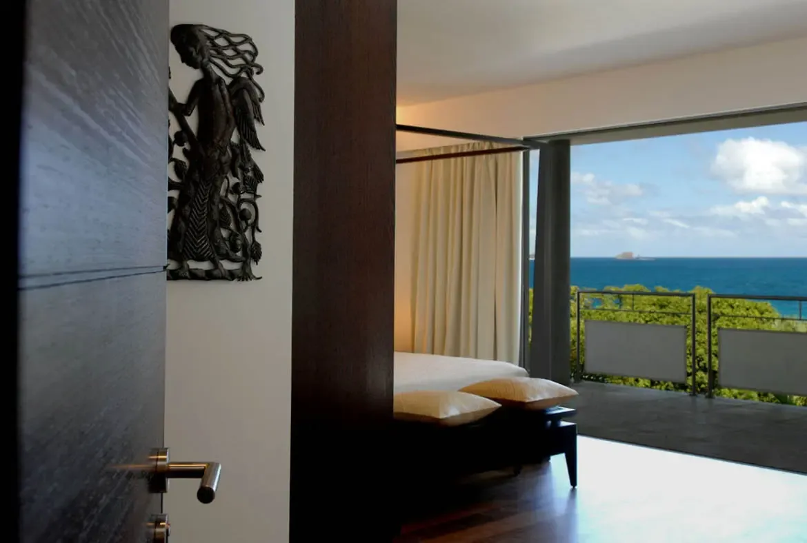 Crisp white linens and sheer curtains frame a bed that faces floor-to-ceiling glass doors opening to an endless blue ocean. A dark wooden wall sculpture adds artistic contrast while lush greenery and a distant island complete this bedroom where the Caribbean is your wallpaper.