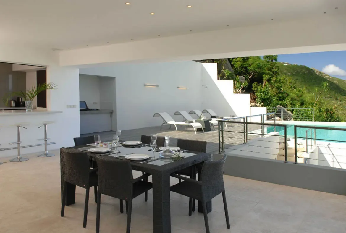 A dark wicker dining table set for six sits on an open-air terrace beside white lounge chairs and the turquoise pool. A sleek bar with silver stools and a kitchen counter nestle under white walls and recessed lighting, while lush green hills roll beneath a bright Caribbean sky—dine, swim, repeat.