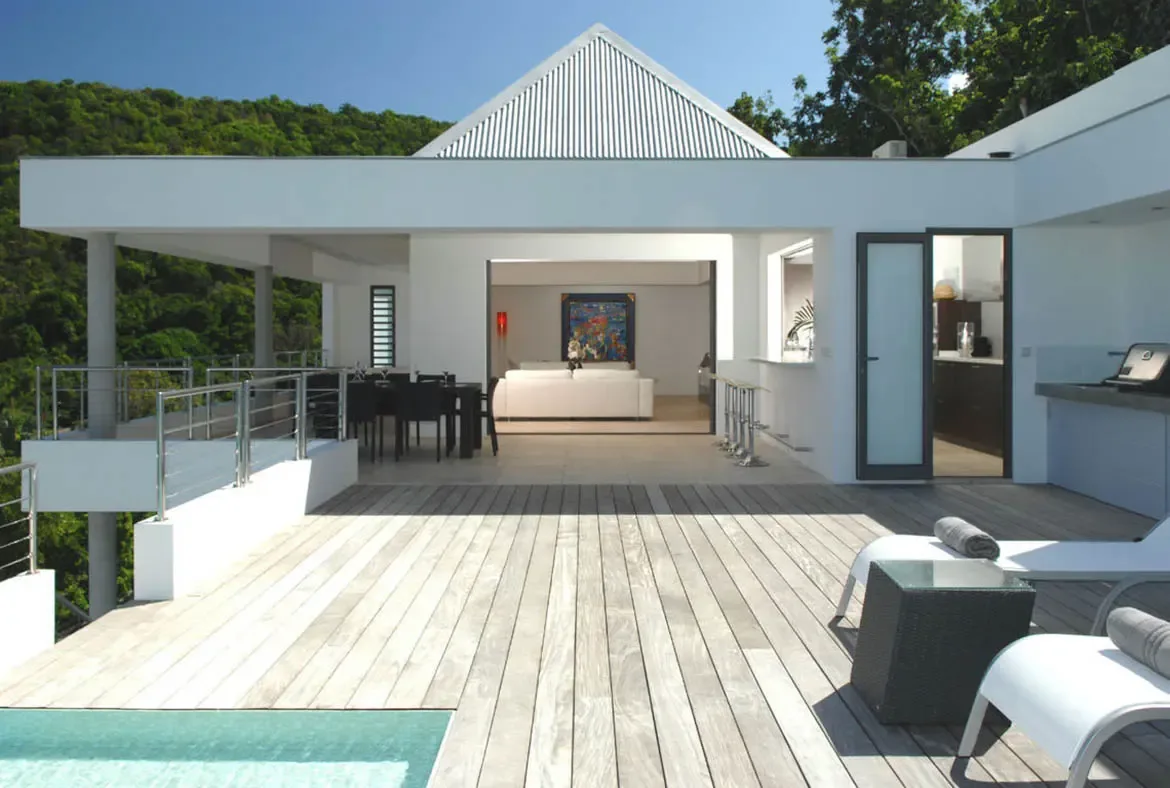 A crisp white gabled villa opens to a spacious wooden deck with lounge chairs and towels beside the turquoise pool. Inside, a colorful abstract painting brightens the white living space while a black dining set sits under the covered terrace—seamless indoor-outdoor Caribbean living.