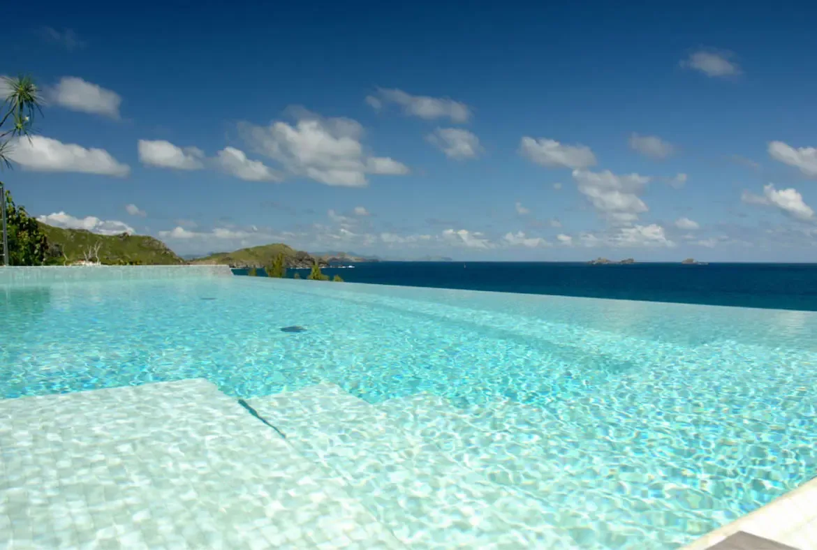 The turquoise infinity pool dissolves into the Caribbean where distant islands rise from deep blue waters. Lush green hills frame the left while palm trees sway overhead—where the pool ends and the ocean begins is anyone's guess.