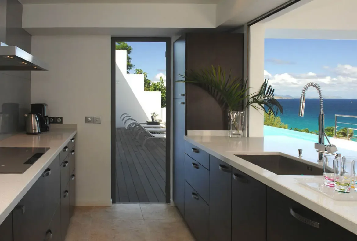 Dark charcoal cabinets and sleek stainless steel appliances define this modern kitchen where a glass vase with a palm frond adds tropical warmth. Sliding glass doors open to a wooden deck with lounge chairs and an infinity pool that merges with the turquoise Caribbean—cooking with an ocean view never looked this good.