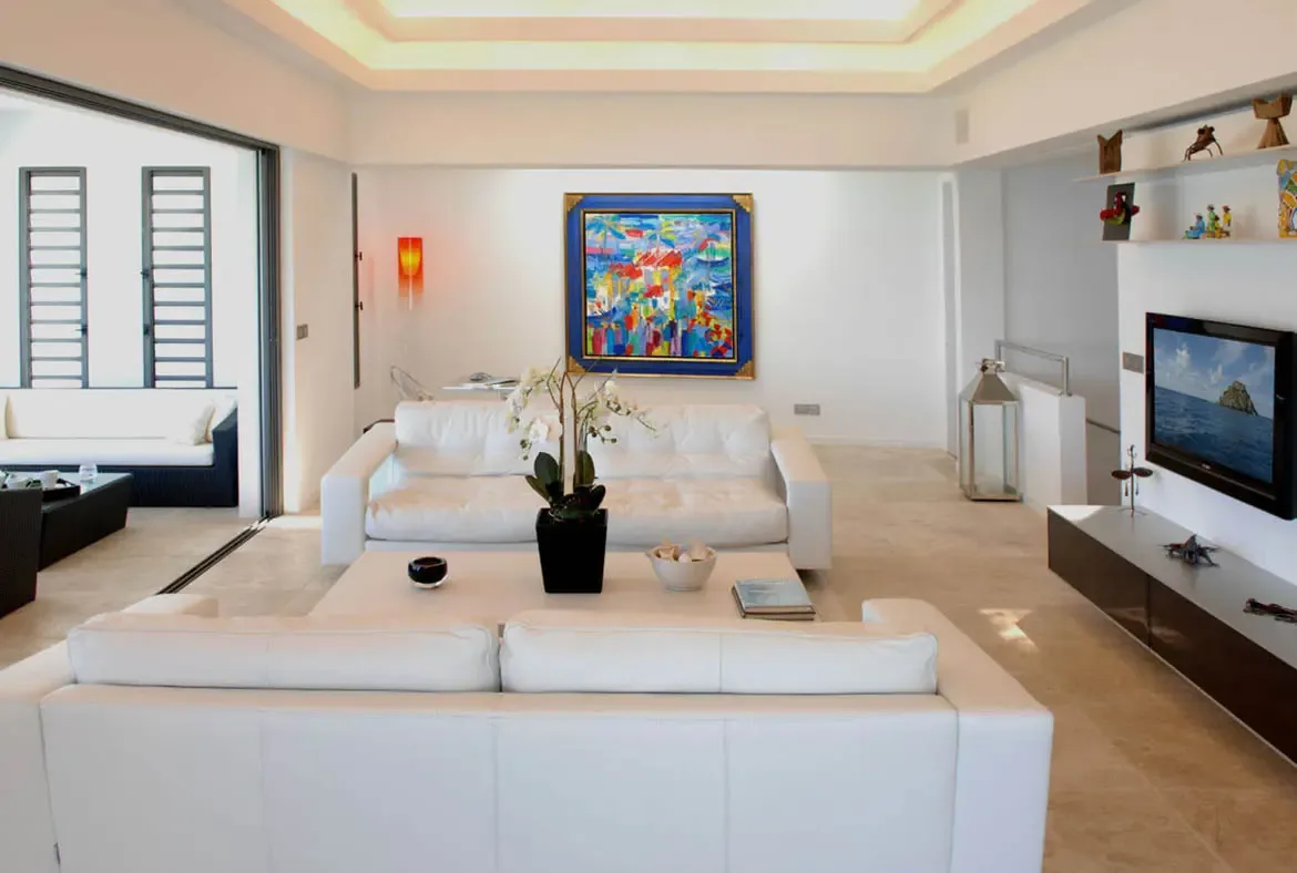 White leather sofas face a flat-screen TV displaying a beach scene, while a vibrant abstract painting in a blue frame and a bold red floor lamp add color to the neutral palette. A black vase of white orchids graces the coffee table—contemporary Caribbean elegance with artistic flair.