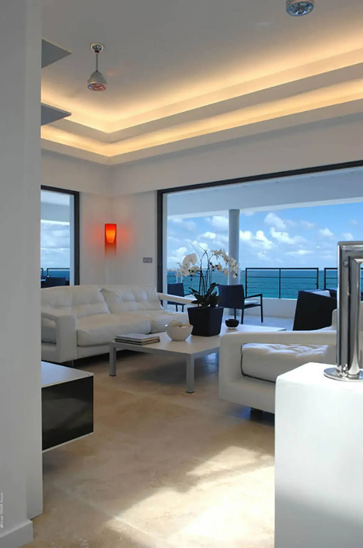 A white sectional sofa and armchair with a low coffee table topped with white orchids in a black planter face floor-to-ceiling glass doors. Beyond the balcony railing, the Caribbean stretches to the horizon—a living room where the ocean is your centerpiece.