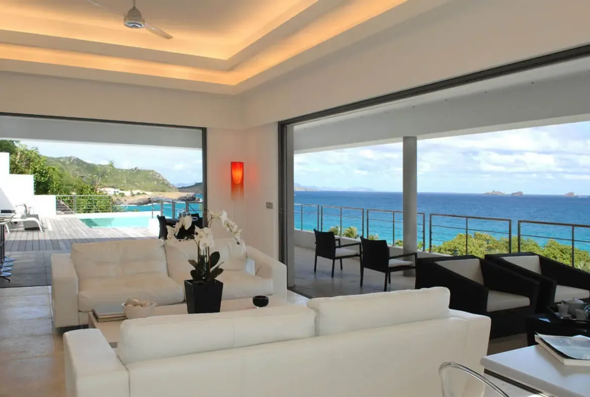 White leather sofas and a coffee table with a black vase of white orchids anchor this sleek living room. Sliding glass doors on both sides open to an infinity pool and the turquoise ocean beyond—indoor-outdoor Caribbean living at its most elegant.
