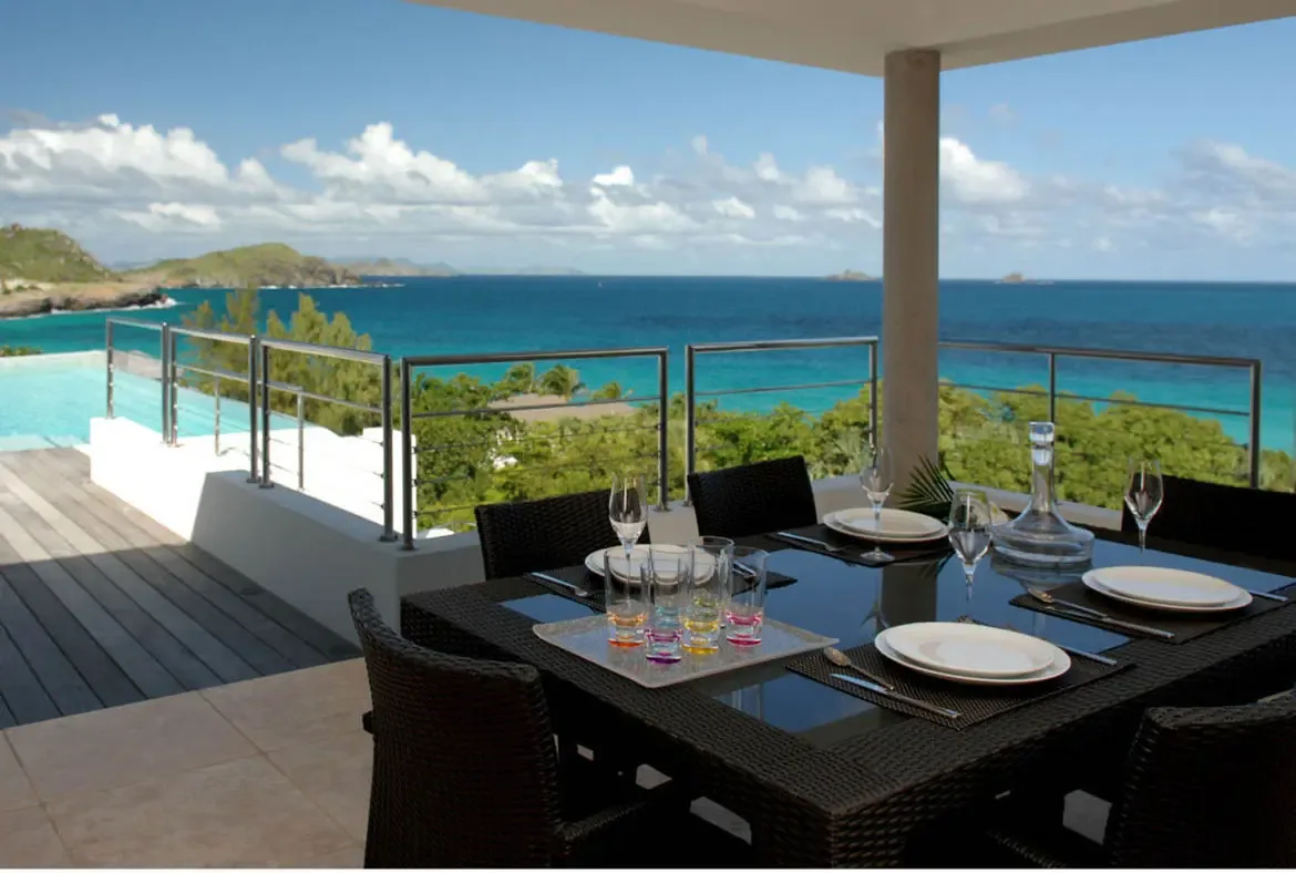 A wicker dining table set for six sits on a tiled patio beside a light blue infinity pool that merges with the Caribbean. Wine glasses and colorful drink tumblers catch the light while distant islands float on turquoise waters—every meal here comes with a million-dollar view.
