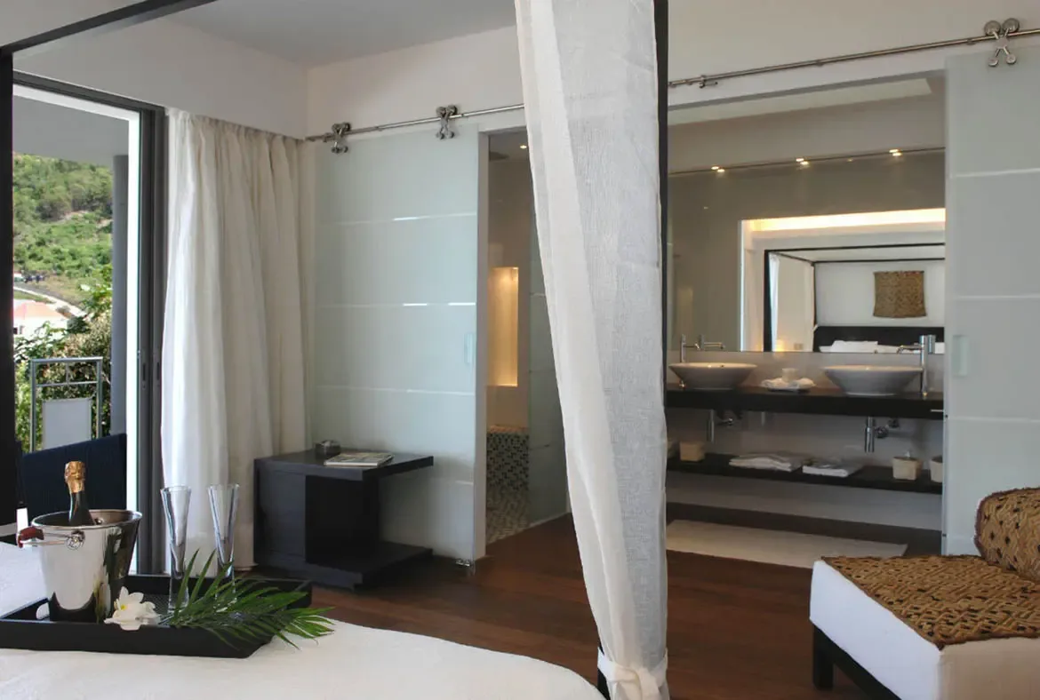 A champagne bottle chills in a silver bucket on the bed beside tall flutes and a white orchid—celebration awaits. The open-concept suite reveals a modern double-vanity bathroom beyond a frosted glass barn door, while sheer curtains frame green hills through the sliding glass doors.