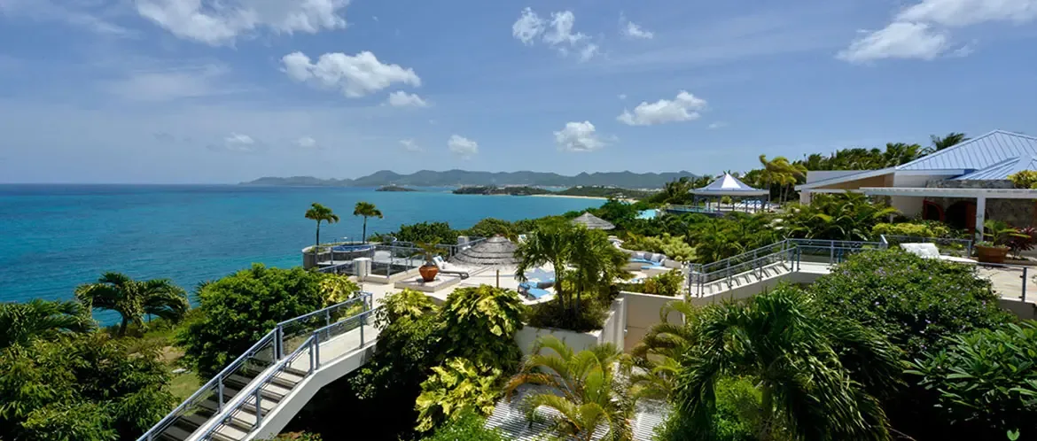 A multi-level white villa with blue roofs clings to a hillside above turquoise ocean, where terraced decks, palapas, and a plunge pool overlook distant islands and a white-sand beach.