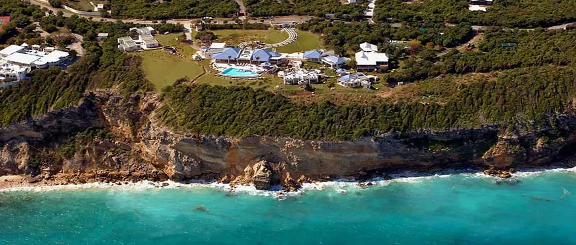 A sprawling white compound with blue roofs perches on rugged cliffs above turquoise water, where manicured lawns and winding paths connect villas overlooking distant islands.