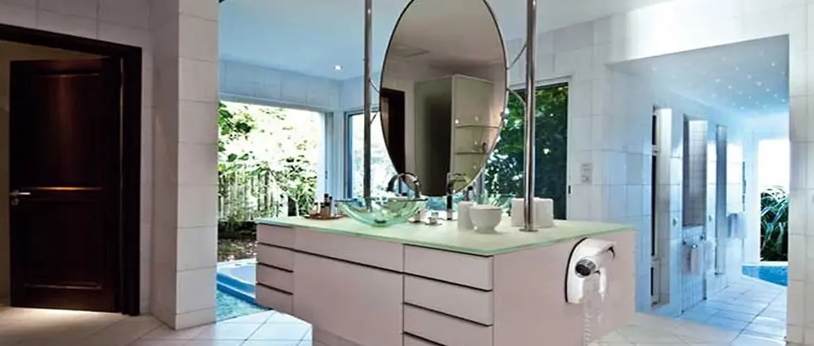 A pale vanity with a green glass vessel sink and chrome faucet sits beneath an oval mirror on chrome poles, where a floor-to-ceiling window reveals lush tropical foliage.