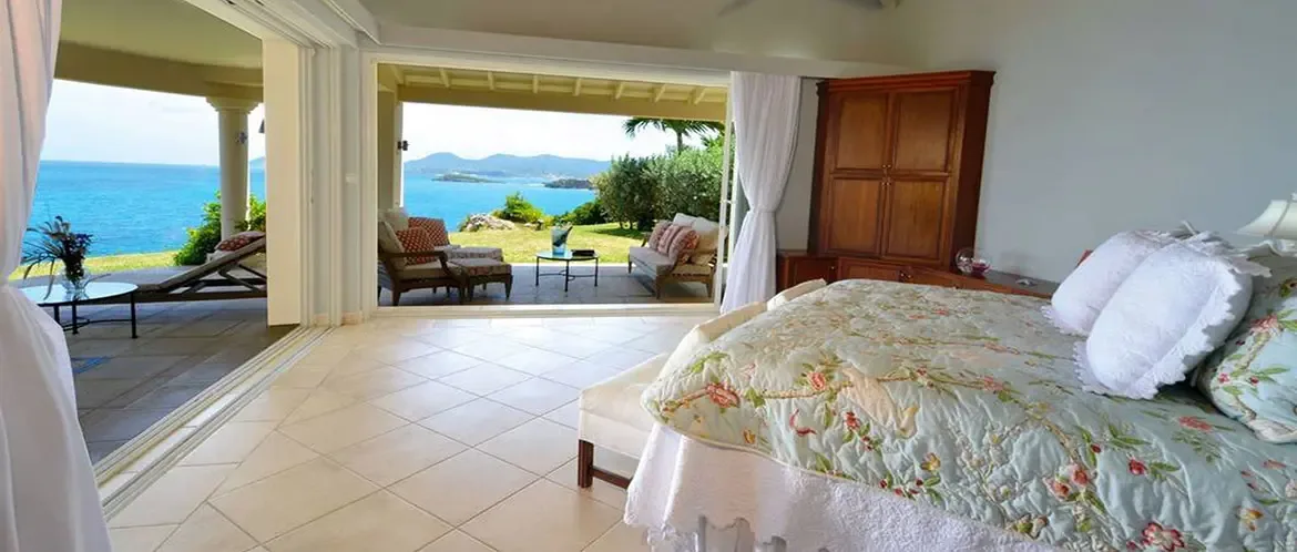 A blue floral quilt covers a bed with white ruffled pillows, where open glass doors lead to a terrace with outdoor chairs and a panoramic turquoise ocean view.