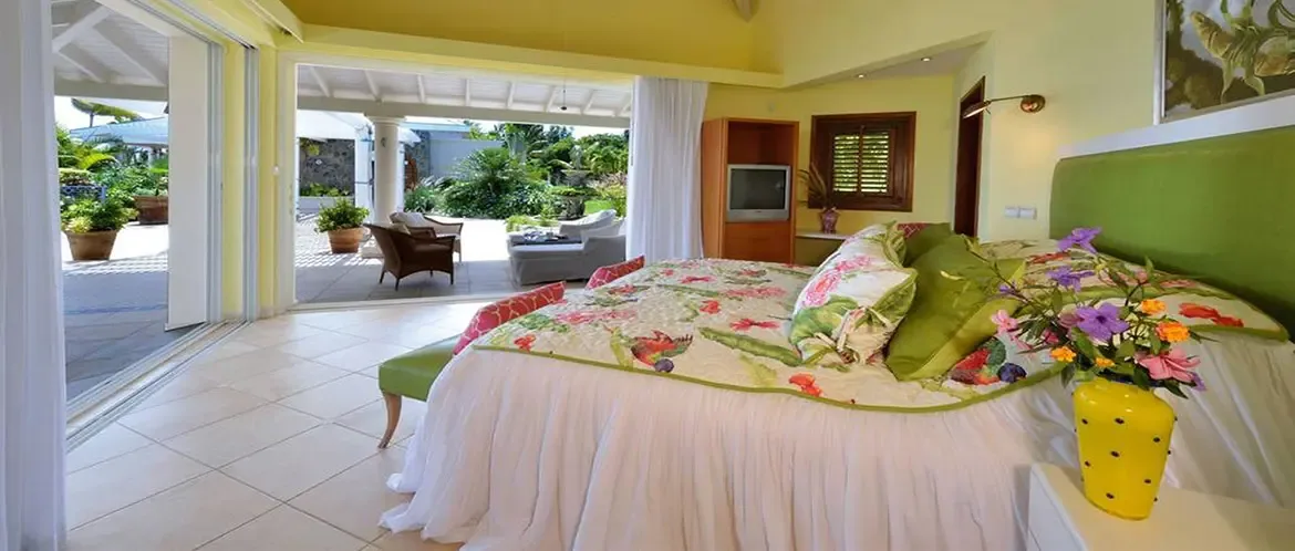A green padded headboard anchors a bed with a tropical floral quilt and lime pillows, where open glass doors lead to a terrace with wicker chairs and an ocean view.