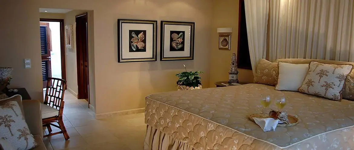A shell-patterned quilt drapes a bed with palm pillows and a tray of white wine, where botanical prints in dark frames hang above and sheer curtains filter the light.