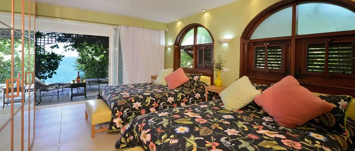 Two beds with navy floral comforters and lime-green pillows sit in a green-walled room, where open glass doors reveal a terrace and a turquoise ocean beyond palm trees.